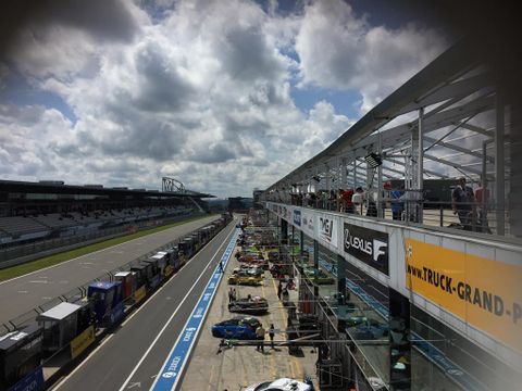Eine Rennstrecke mit Boxengasse und mehreren Rennwagen. Der Himmel ist teils bewölkt mit einigen blauen Stellen.