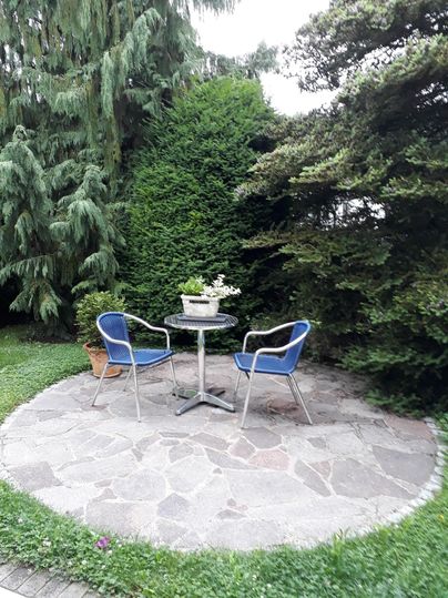 Un petit coin dans le jardin avec une table ronde et deux chaises bleues. Entouré d'arbres et de buissons verts et hauts.
