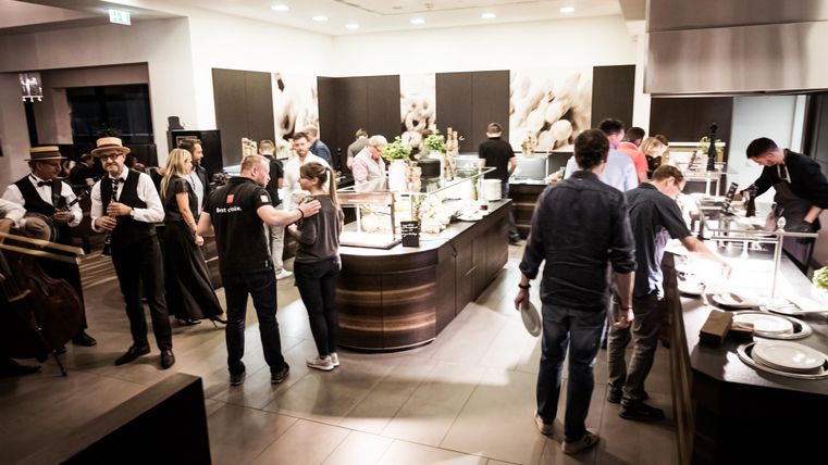 A bustling restaurant kitchen with guests and chefs. There are several stations for food and a pleasant atmosphere.