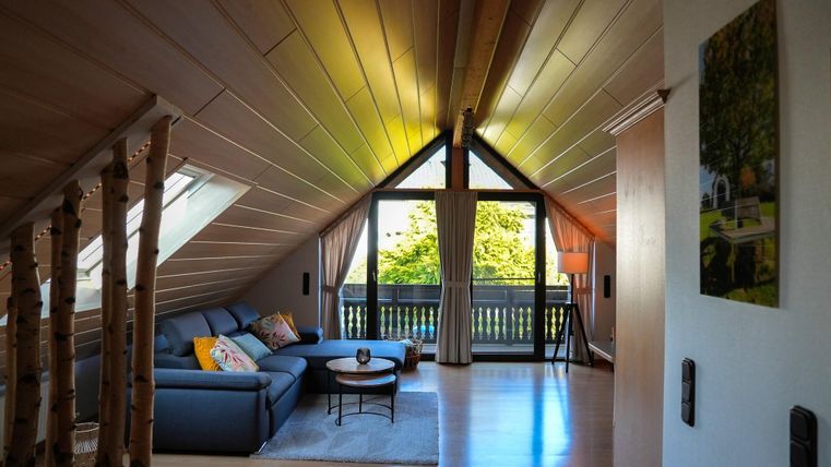 Ein gemütliches Wohnzimmer in einem Dachgeschoss mit Holzdecke. Große Fenster bieten einen Blick auf den Außenbereich und lassen viel Tageslicht herein.