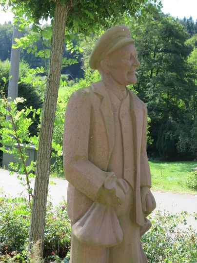 Steinfigur eines Mannes mit Mütze und Mantel in einer grünen Umgebung.