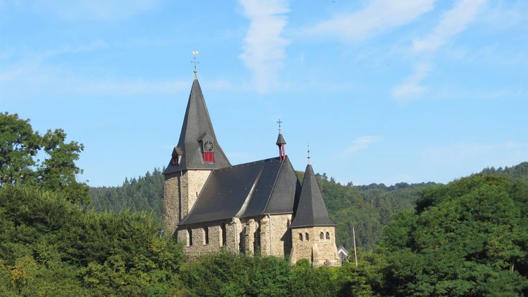 An old church surrounded by trees and hills. The sky is clear and blue with a few clouds.