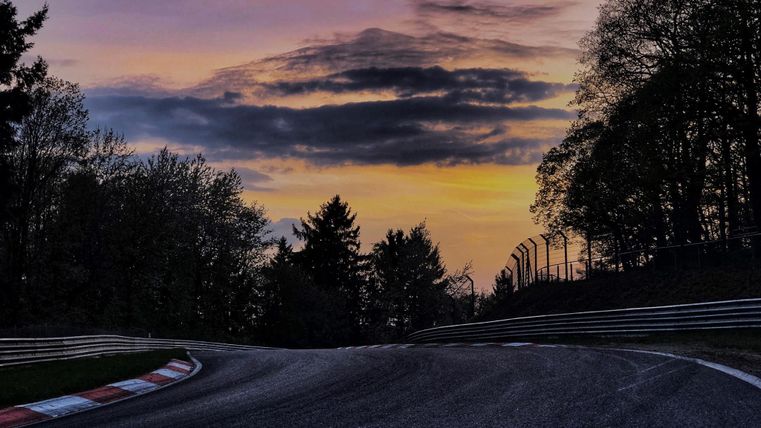 Eine kurvenreiche Rennstrecke unter einem bunten Abendhimmel. Bäume umrahmen die Strecke und die Wolken reflektieren das Licht des Sonnenuntergangs.