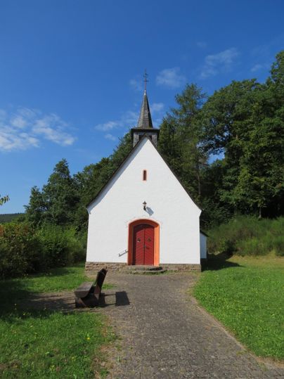 Eine kleine weiße Kapelle steht in einer grünen Umgebung. Im Vordergrund sind Bänke und ein klarer Himmel sichtbar.