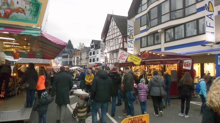 Eine belebte Straße mit vielen Menschen, die an Ständen entlangschlendern. Im Hintergrund sind typische Fachwerkhäuser und bunte Verkaufsstände zu sehen.