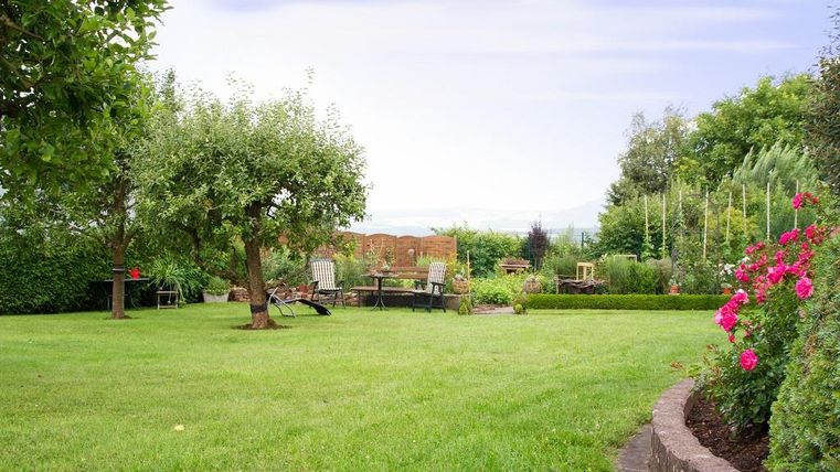 A well-maintained garden with green grass, fruit trees, and colorful flowers. In the background, garden furniture and a tranquil landscape are visible.