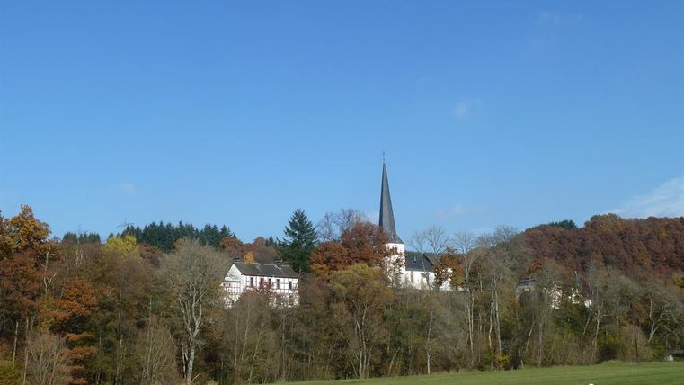 A picturesque village with a church and a tall tower amidst autumn-colored trees. The sky is clear and blue, while the meadow in the foreground shines green.