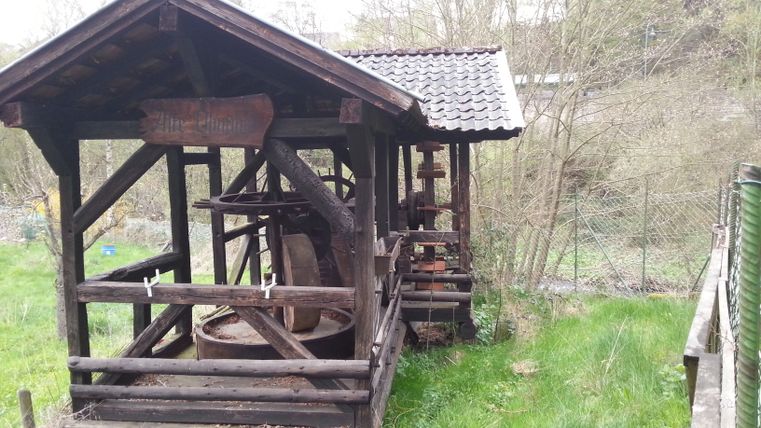 Alte hölzerne Ölmühle mit Dach in einer grünen Umgebung.