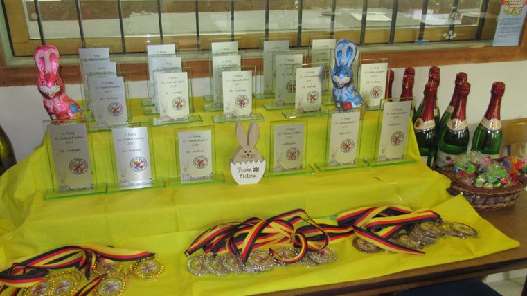 Eine Tischdeko mit Preisen, Medaillen und Schokoladenhasen. Die Auszeichnungen sind auf einem gelben Tischdecke angeordnet, umgeben von Sektflaschen.