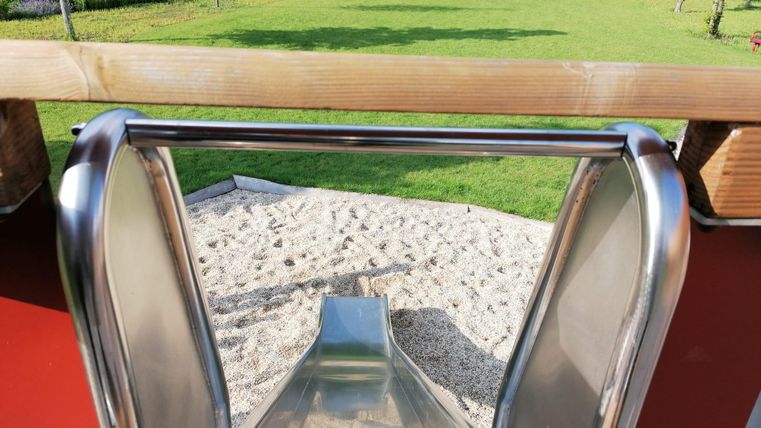 Eine Rutsche in einem Spielplatz mit Blick auf den Sandbereich. Im Hintergrund sind grüne Bäume und eine gepflegte Wiese zu sehen.