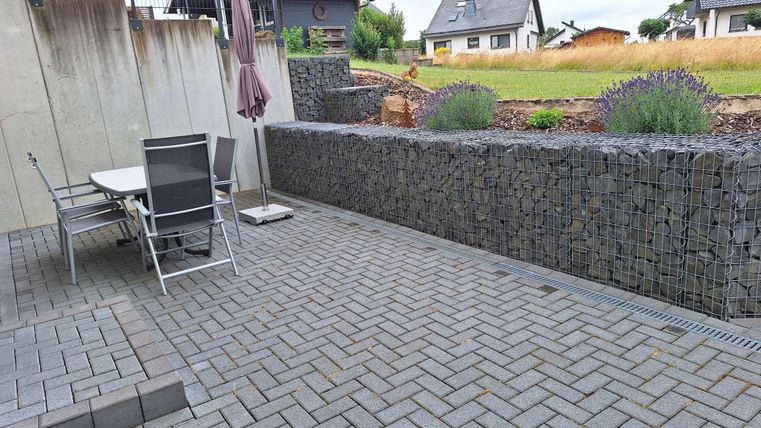Un patio moderne avec des pavés gris et une table de jardin avec des chaises. À l'arrière-plan, on peut voir quelques maisons et un champ cultivé.