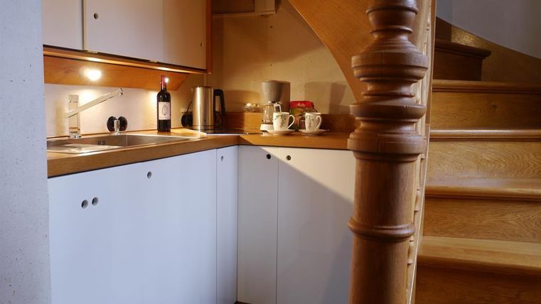 A modern kitchen with wooden shelves and a wooden staircase. On the countertop, there are cups and a bottle of wine.