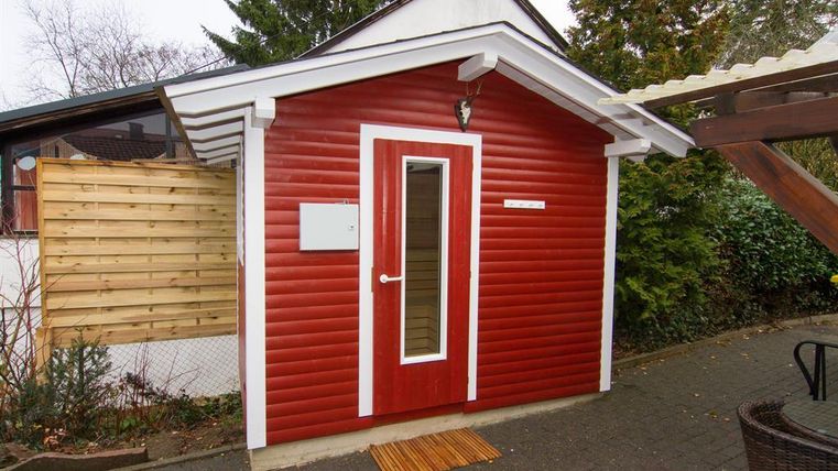 A small red house with a white door stands outside. Surrounded by trees and plants.