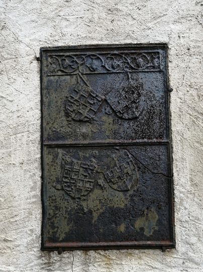 An old, weathered plaque with two coats of arms. The plaque is located on a rough, light wall.