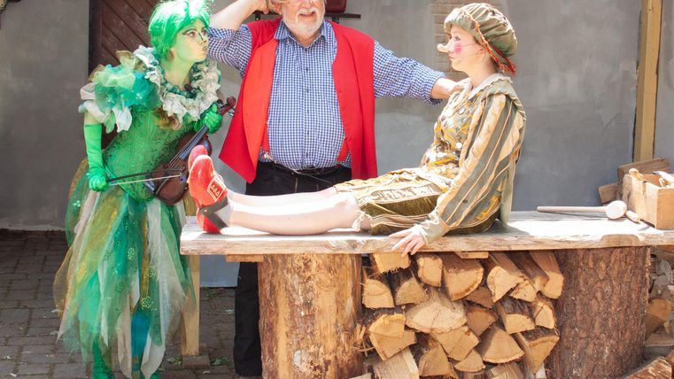 Ein bunter Auftritt mit drei Darstellern in fantasievollen Kostümen. Sie stehen an einem Holzstapel und scheinen eine lebhafte Szene zu spielen.