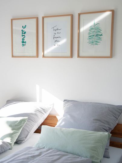 Ein gemütliches Schlafzimmer mit hellen Kissen und drei gerahmten Bildern an der Wand. Die Sonne scheint sanft auf das Bett.