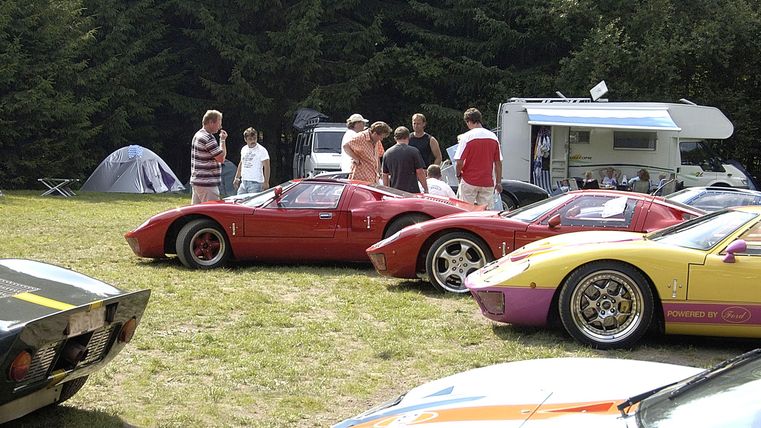 A group of people is standing around several classic cars on a meadow. In the background, a tent and trees can be seen.