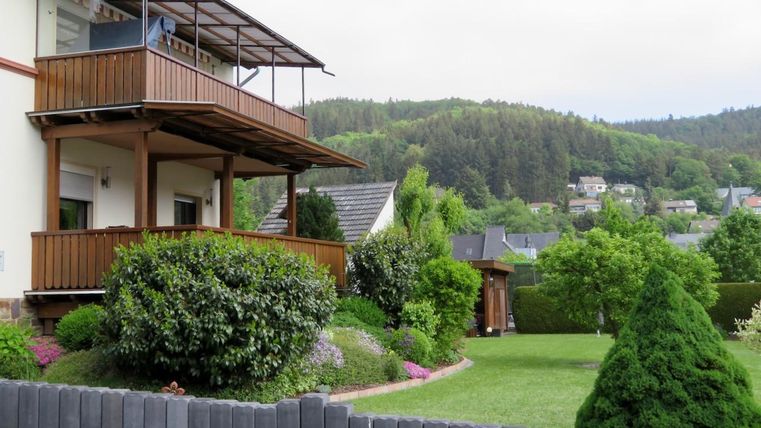 Une maison moderne avec un balcon et un jardin vert. En arrière-plan, on peut voir des collines douces et d'autres maisons résidentielles.