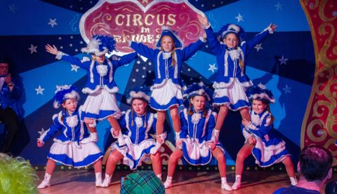 Een groep danseressen in blauwe kostuums voert een vrolijke performance op. Op de achtergrond is een kleurrijk circus-thema te zien.