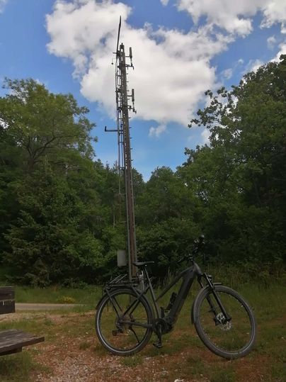 Een elektrische fiets staat naast een hoge zendmast in een bosgebied. De lucht is helder met enkele wolken.
