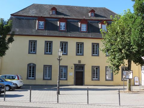 Ein historisches Gebäude mit gelber Fassade und mehreren Fenstern. Im Vordergrund sind Bäume und ein Parkplatz zu sehen.