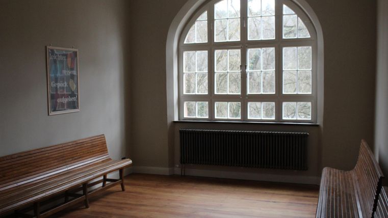 Lege wachtruimte met houten banken, groot raam en een poster aan de muur. Houten vloer en radiator onder het raam.