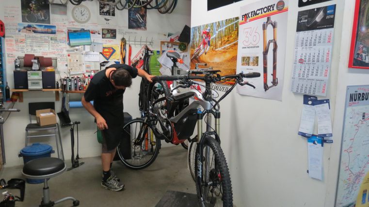 Ein Mechaniker arbeitet an einem Fahrrad in einer Werkstatt. Im Hintergrund hängen weitere Fahrräder und verschiedenen Werkzeuge sind sichtbar.