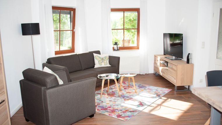 Ein gemütliches Wohnzimmer mit einem braunen Sofa und einem Kleinen Tisch. Große Fenster lassen viel Licht herein und der Fernseher steht auf einem modernen Möbelstück.