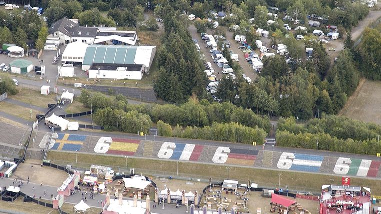 An aerial view of a racetrack with colorful markings. In the background, camping areas and green spaces can be seen.