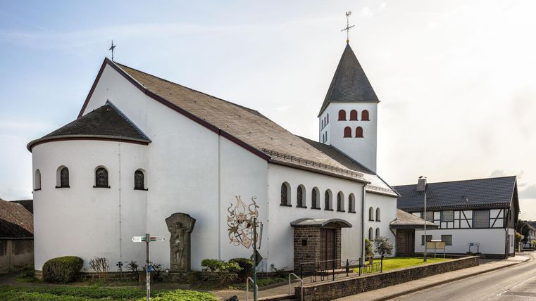 Eine weiße Kirche mit einem charakteristischen Turm steht an einer Straße. Der umgebende Garten ist gepflegt und es sind einige Gebäude in der Nähe sichtbar.