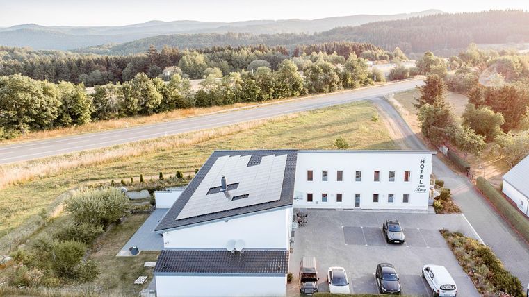 Een modern gebouw met zonnepanelen op het dak, omringd door groene bomen. Ernaast loopt een weg waar parkeerplaatsen zijn.