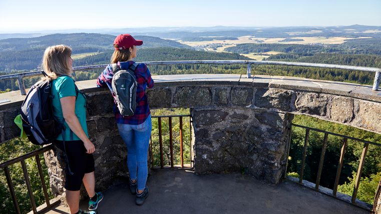 Zwei Personen stehen auf einem Aussichtsturm und blicken in die Ferne über eine bewaldete Landschaft.