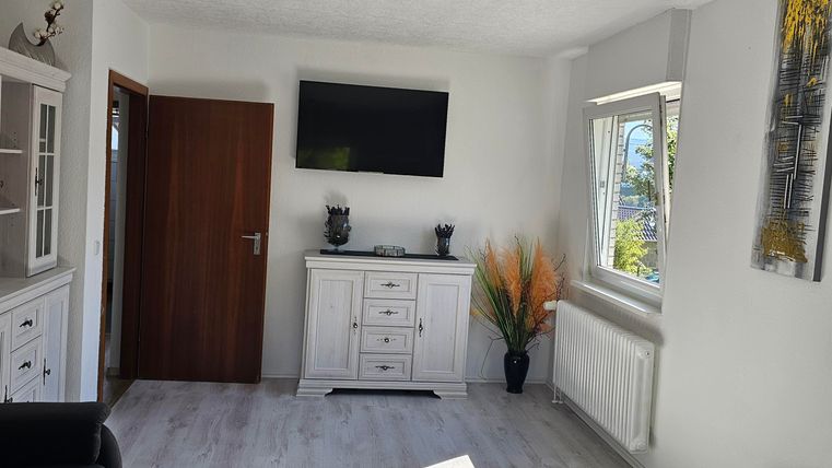 A modern living room with a white sideboard, a television on the wall, and a small table. The atmosphere is bright and welcoming, with a plant arrangement in the corner.