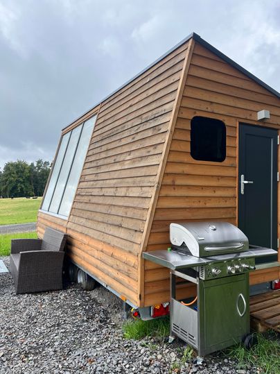 Une maison tiny moderne avec une façade en bois et de grandes fenêtres. Dehors se trouve un grill, et l'environnement est composé de prairies vertes.