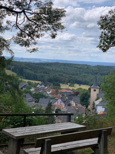 Ein Blick auf ein malerisches Dorf umgeben von grüner Landschaft. Im Vordergrund steht eine Holzbank unter einem Baum.