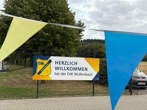 Ein Willkommensschild der DJK Müllenbach, umgeben von bunten Fahnen. Die Umgebung ist grün und ländlich.
