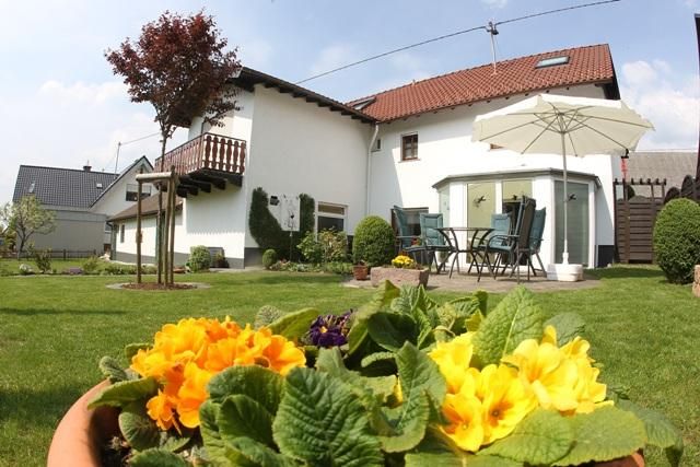 Een gezellig huis met een verzorgde tuin. Kleurrijke bloemen bloeien op de voorgrond, en een tafel met stoelen staat op het terras.