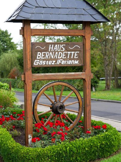 A wooden sign with the inscription "Haus Bernadette" and a wagon wheel. Surrounded by colorful flowers and bushes in the garden.