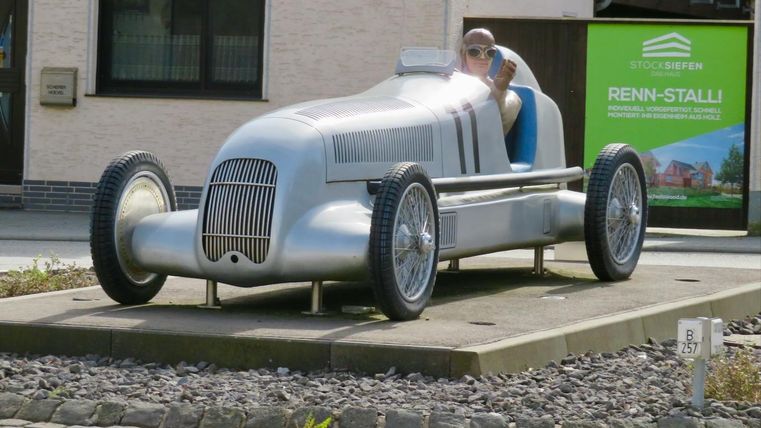 Une voiture de course vintage argentée sur un socle. À l'intérieur de la voiture, une personne avec des lunettes de soleil est assise.