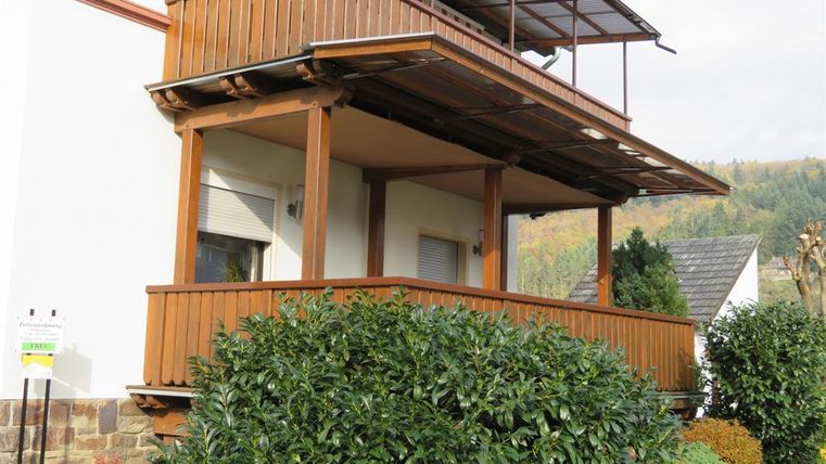 Une maison moderne avec un balcon en bois. L'environnement est aménagé avec des plantes vertes.