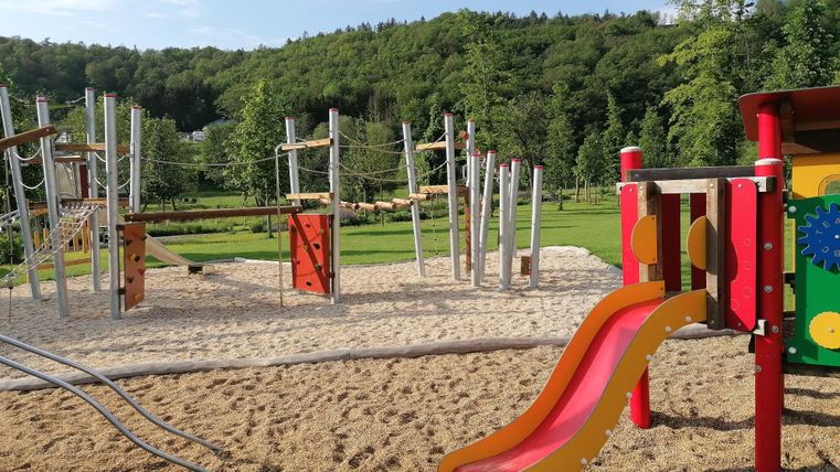 Ein Spielplatz mit einer Rutsche und Klettergerüsten inmitten einer grünen Landschaft. Der Sandboden lädt zum Spielen ein.