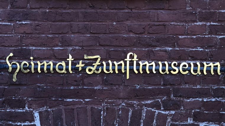 Ein Schild mit der Aufschrift "Heimat + Zunftmuseum" an einer roten Backsteinmauer. Die goldene Schrift hebt sich gut vom Hintergrund ab.