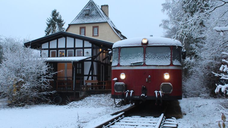A red, snow-covered train stands on the tracks next to a building in a snowy landscape. The scene is calm and peaceful, snow covers the ground and the trees.