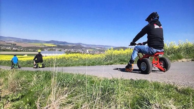 Drie personen rijden op kleine quads op een pad. Op de achtergrond zijn gele koolzaadvelden en een glooiend landschap te zien.