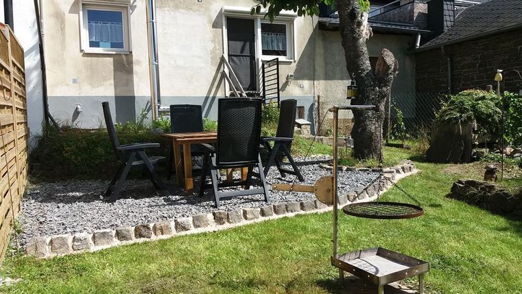 Ein gemütlicher Garten mit einem Essbereich aus Holz und schwarzen Stühlen. Im Hintergrund steht ein Grill und es gibt viel Grünfläche.