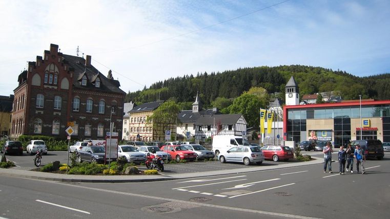 Un paysage urbain animé avec plusieurs véhicules et piétons. À l'arrière-plan, une colline boisée et divers bâtiments sont visibles.