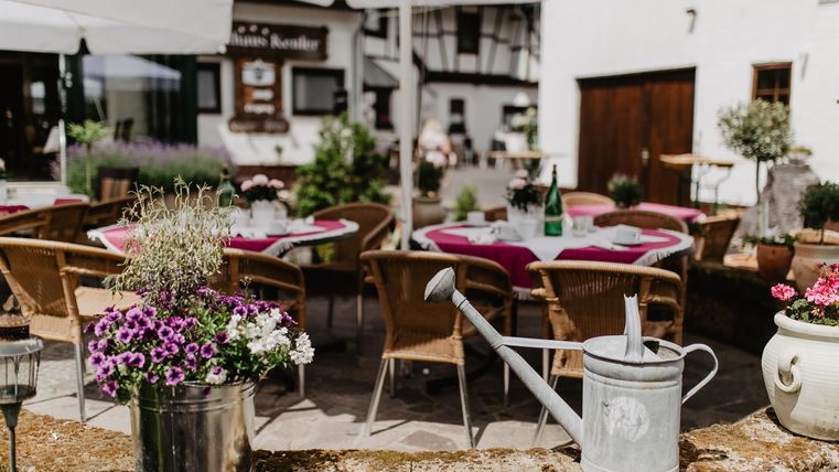 Ein gemütlicher Außenbereich mit runden Tischen und Stühlen, umgeben von Blumen. Sonnenschirme schützen die Gäste vor der Sonne.