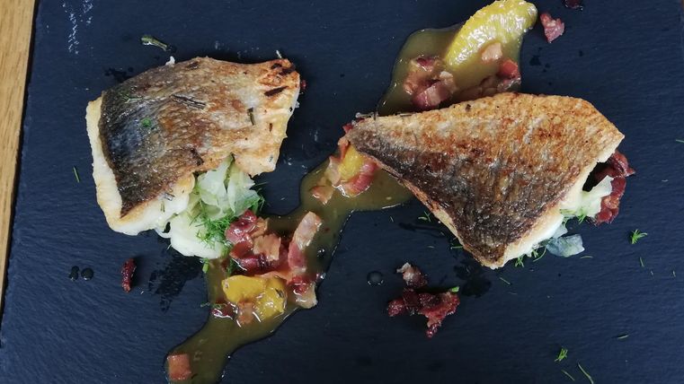 Een gerecht met gebakken vis op een zwart bord. Het is gegarneerd met groenten en een saus.