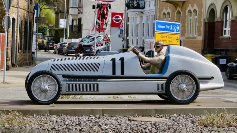 Une voiture historique avec le numéro 11 roule sur une route. Le conducteur est habillé d'une tenue nostalgique et apprécie la balade.