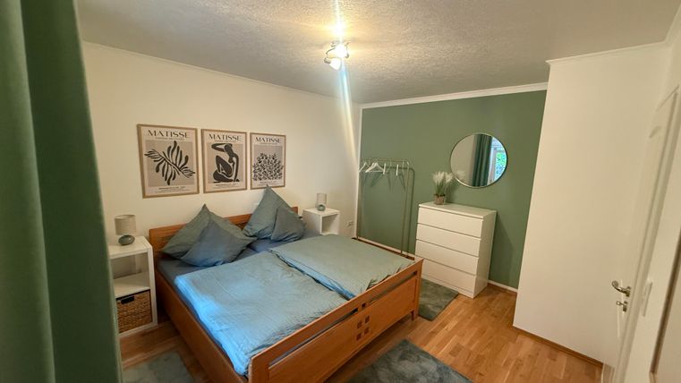 Een gezellige slaapkamer met een tweepersoonsbed en minimalistische inrichting. De muren zijn in zachte groentinten geschilderd, en er zijn decoratieve schilderijen en een grote spiegel.